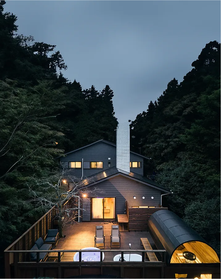 The villa at dusk, its lit deck and barrel sauna set against the forest