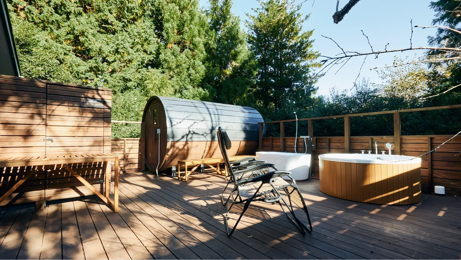 Barrel sauna and round jacuzzi on the sunlit deck, ringed by trees